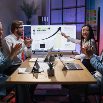 Asian woman presenting financial report of worldwide economy on big monitor during meeting with international partners. Diverse colleagues sitting at desk and appealing to her speech.